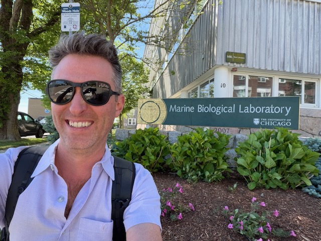 Dr. Jason Gallant smiling in front of the Marine Biological Laboratory sign in Woods Hole, Massachusetts